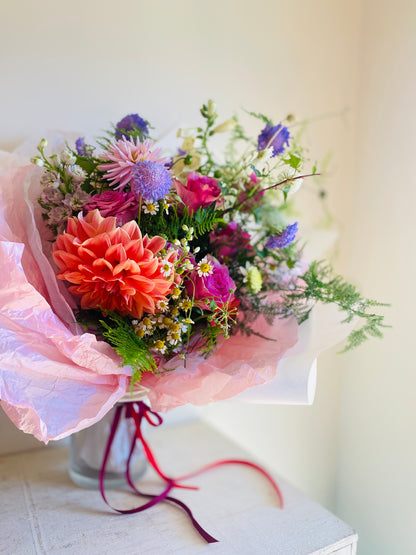 A Valentine's Day floral arrangement featuring a variety of colorful flowers in pink and red tones, wrapped in pink and white paper with pink and red ribbons.